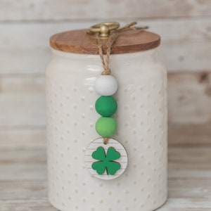 May include: A cream-colored jar with a wooden lid and a decorative beaded charm. The charm features white and green beads, and a wooden disc with a green four-leaf clover design. The jar has a dotted pattern.