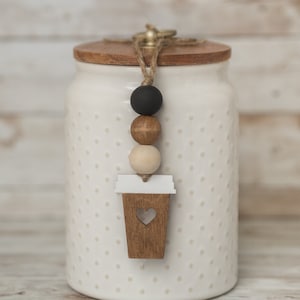 May include: White ceramic jar with a wooden lid and a brown and white coffee cup charm hanging from the lid. The jar has a white polka dot pattern.