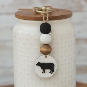 May include: White ceramic canister with a wooden lid and a black and white cow silhouette charm. The charm is attached to a string with black, white, and gold beads. The canister is sitting on a wooden cutting board.