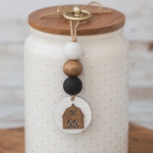 May include: White ceramic canister with a wooden lid and a farmhouse-style wooden charm hanging from the lid. The charm features a small wooden house with a red roof and white accents. The canister has a white polka dot pattern.