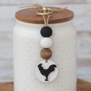 May include: White ceramic canister with a wooden lid and a black rooster charm. The canister has a polka dot pattern and is sitting on a wooden cutting board.