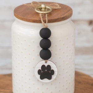 May include: White ceramic jar with a wooden lid and a black paw print charm. The jar has a polka dot pattern and a gold knob on the lid. The charm is hanging from a brown string with black beads.