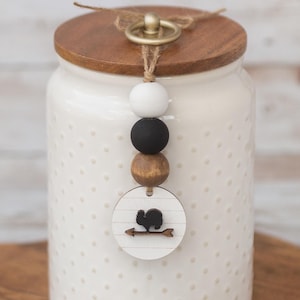 May include: A white ceramic jar with a wooden lid and a decorative bead strand. The jar has a dotted pattern and a wooden tag with a squirrel and arrow design. The jar is on a wooden cutting board.