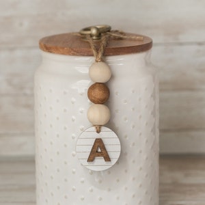 May include: White ceramic jar with a wooden lid and a wooden bead garland with a letter A charm. The jar has a dotted pattern.