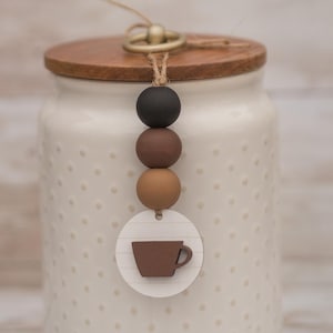May include: A cream-colored ceramic jar with a wooden lid and a decorative tag. The tag features a brown coffee cup design and is attached to a string of black, brown, and tan beads. The jar has a dotted pattern.