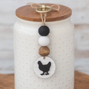 May include: White ceramic canister with a wooden lid and a black and white chicken silhouette charm. The canister has a polka dot pattern and is sitting on a wooden cutting board.