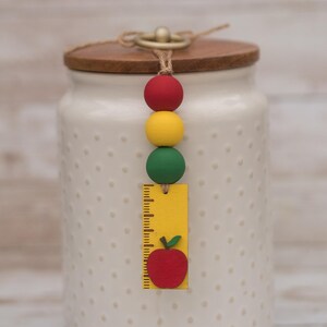May include: A white ceramic jar with a wooden lid and a decorative beaded charm. The charm features red, yellow, and green beads, and a yellow ruler-shaped tag with a red apple illustration. The jar has a dotted pattern.
