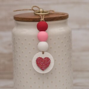 May include: A white ceramic jar with a wooden lid and a beaded charm. The charm features a red, pink, and white bead stack, and a small round tag with a glittery red heart. The jar has a dotted pattern.