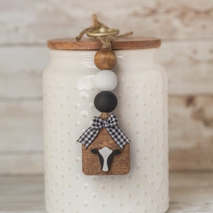 May include: White ceramic jar with a wooden lid and a black and white cow charm hanging from the lid. The jar has a polka dot pattern and the charm has a black and white checkered bow.