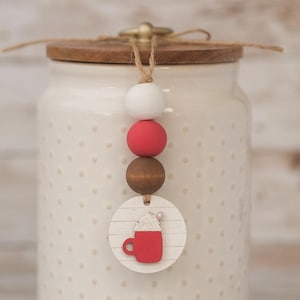 May include: A cream-colored ceramic jar with a wooden lid and a decorative beaded accent. The accent features a white, red, and brown bead design, and a wooden tag with a red mug graphic. The jar has a dotted pattern.