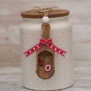 May include: A white ceramic jar with a wooden lid and a decorative wooden scoop. The scoop is adorned with a red ribbon featuring white hearts and the letters "XO". The jar has a dotted pattern.