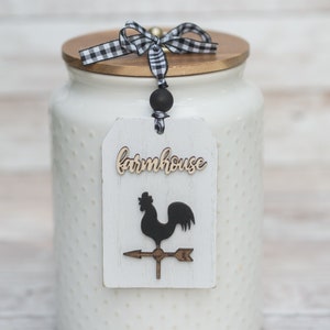 May include: A white ceramic jar with a wooden lid, adorned with a black and white checkered ribbon bow. A white tag with the word "farmhouse" and a rooster weathervane design hangs from the jar. The jar has a dotted texture.