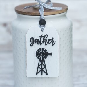May include: A white ceramic jar with a wooden lid and a tag that says "gather". The tag has a black windmill design. A gray ribbon is tied around the lid. The jar has a dotted pattern.