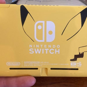 Könnte beinhalten: Gelbe Nintendo Switch Konsole mit Pikachu-Design. Die Konsole hat ein weißes Nintendo Switch Logo und den Text "NINTENDO SWITCH" in weiß.