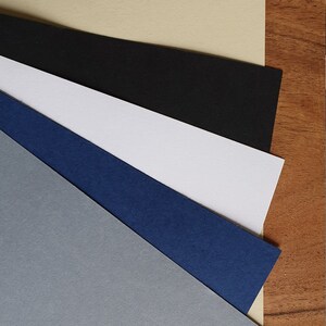 Op de afbeelding: Vijf vellen papier in verschillende kleuren, van boven naar beneden: beige, zwart, wit, blauw en grijs.