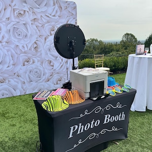 May include: A photo booth setup outdoors with a backdrop of white roses. The black table cover reads "Photo Booth" in white script. Accessories include novelty hats and sunglasses. A large ring light and printer are also visible.