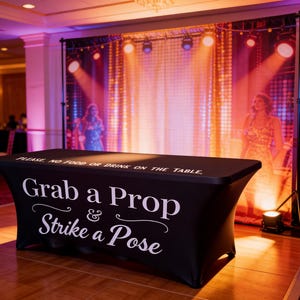 May include: A black table with the text "Grab a Prop & Strike a Pose" in white script. The table is in front of a backdrop with a singer and stage lights. The backdrop is supported by a black stand.
