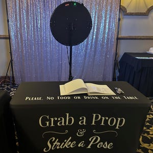 May include: A photo booth setup with a black table cover that reads "Grab a Prop & Strike a Pose." A large, round black light is positioned above the table, in front of a shimmering, sequined backdrop. A sign on the table says "PLEASE. NO FOOD OR DRINK ON THE TABLE."