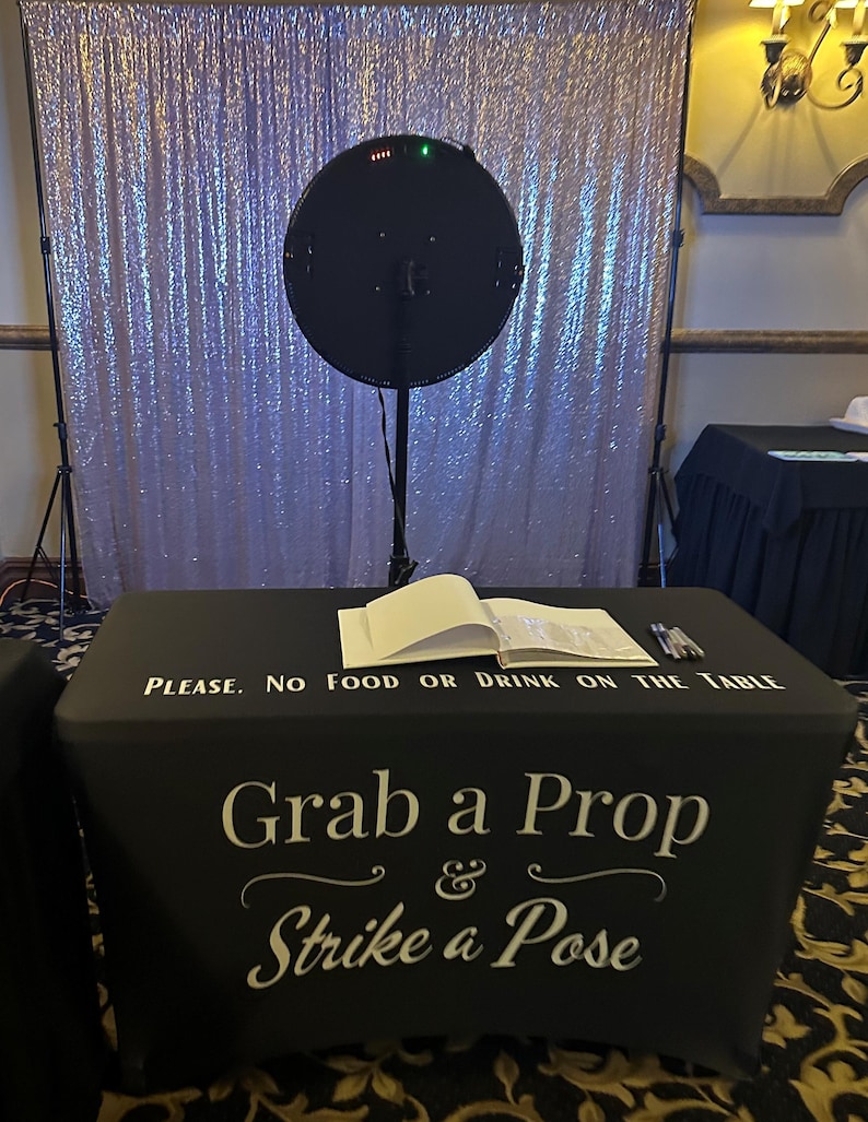 May include: A photo booth setup with a black table cover that reads "Grab a Prop & Strike a Pose." A large, round black light is positioned above the table, in front of a shimmering, sequined backdrop. A sign on the table says "PLEASE. NO FOOD OR DRINK ON THE TABLE."
