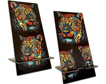 Puede incluir: Dos soportes rectangulares de acrílico negro con un diseño de estampado de leopardo vibrante y multicolor. Cada soporte tiene una base plana y una pequeña repisa horizontal para sostener objetos, decorada con ilustraciones estilizadas de cabezas de leopardo.