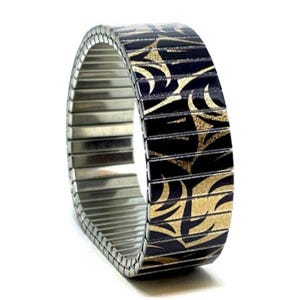Puede incluir: Una pulsera de reloj de acero inoxidable con estampado negro y dorado. La pulsera está hecha de eslabones metálicos entrelazados y tiene un diseño flexible.