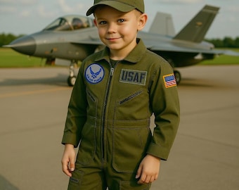 May include: A young child wearing an olive green USAF flight suit and matching cap with an American flag patch. The suit has a US Air Force patch and an American flag patch. A military jet is in the background.