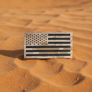 Puede incluir: Un parche rectangular con el diseño de la bandera estadounidense. Las estrellas y las rayas están representadas en una paleta de colores apagados, inspirada en el desierto, con un aspecto texturizado. El parche se encuentra sobre un fondo de dunas de arena.