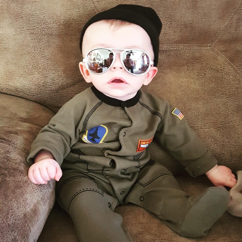 Puede incluir: Un beb&eacute; con gorro negro, gafas de aviador y un mono verde. El mono tiene parches con un avi&oacute;n y la bandera estadounidense. Las gafas reflejan la habitaci&oacute;n. El beb&eacute; est&aacute; sentado en un sof&aacute; marr&oacute;n.