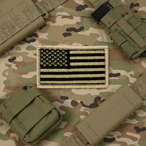 May include: A rectangular patch featuring the American flag in black and tan. The flag design includes stars in the upper left corner and horizontal stripes. The patch is surrounded by a tan border and is set against a camouflage background.