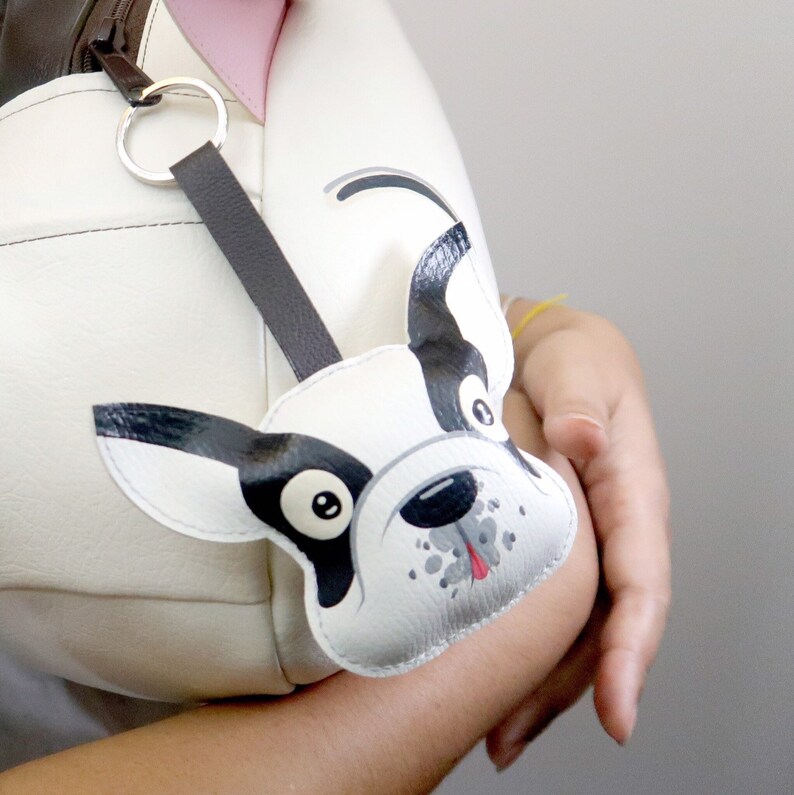 K&ouml;nnte beinhalten: Eine wei&szlig;e Ledertasche mit einem schwarz-wei&szlig;en Cartoon-Hundeanh&auml;nger an einem braunen Riemen. Der Hund hat schwarze Ohren, eine schwarze Nase und eine rosa Zunge.