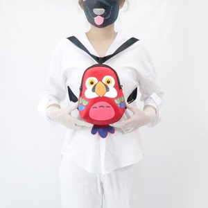 May include: A red parrot-shaped backpack with colorful details. The backpack has a black adjustable strap and a zipper closure.