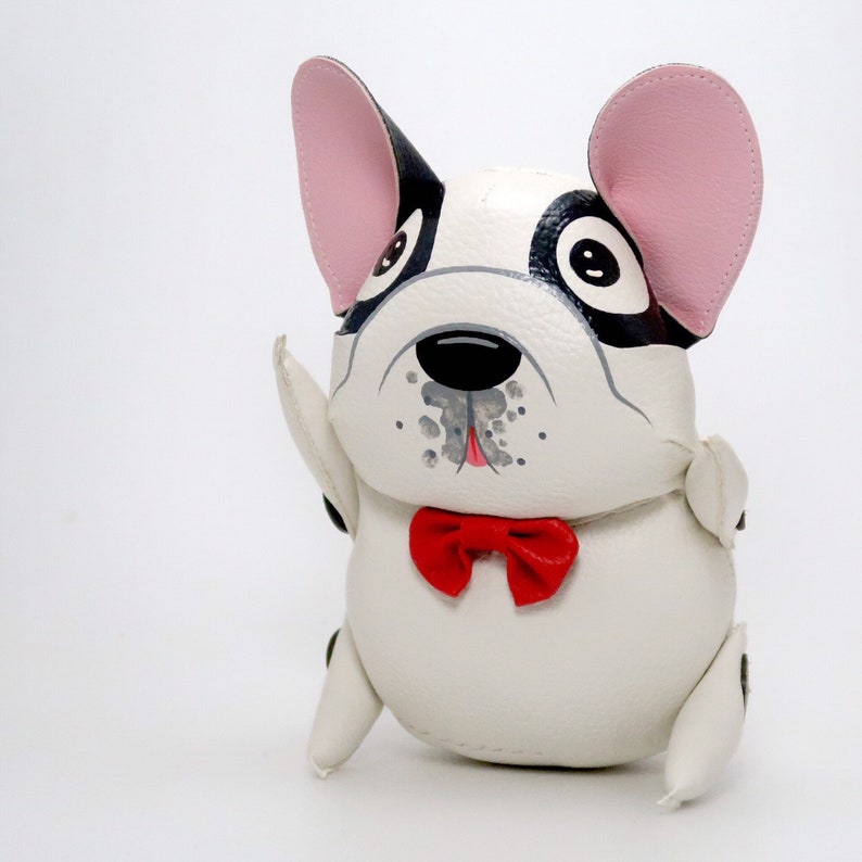 K&ouml;nnte beinhalten: Ein wei&szlig;es Pl&uuml;schtier in Form eines Hundes mit schwarzen und grauen bemalten Details, rosa Ohren und einer roten Schleife. Der Hund sitzt mit erhobenen Vorderpfoten.