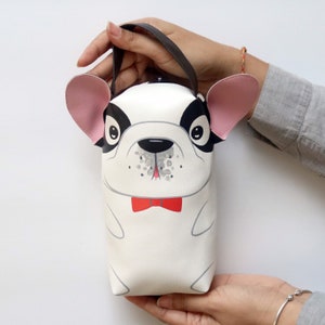K&ouml;nnte beinhalten: Eine wei&szlig;e und schwarze Handtasche in Form einer Franz&ouml;sischen Bulldogge mit rosa Ohren und einer roten Schleife. Die Tasche hat einen schwarzen Henkel.