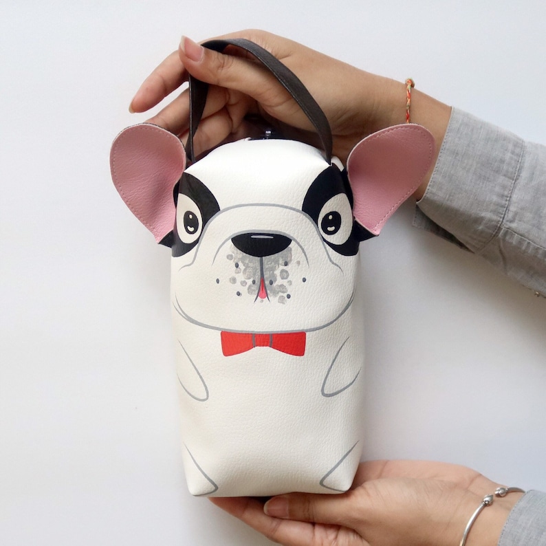K&ouml;nnte beinhalten: Eine wei&szlig;e und schwarze Handtasche in Form einer Franz&ouml;sischen Bulldogge mit rosa Ohren und einer roten Schleife. Die Tasche hat einen schwarzen Henkel.