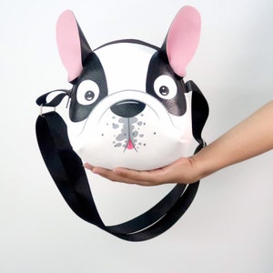 K&ouml;nnte beinhalten: Eine schwarze und wei&szlig;e Umh&auml;ngetasche in Form eines franz&ouml;sischen Bulldoggen mit rosa Ohren und einem schwarzen verstellbaren Riemen.