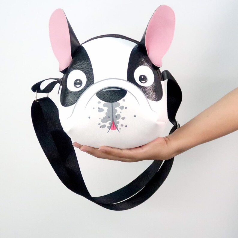 K&ouml;nnte beinhalten: Eine schwarze und wei&szlig;e Umh&auml;ngetasche in Form eines franz&ouml;sischen Bulldoggen mit rosa Ohren und einem schwarzen verstellbaren Riemen.