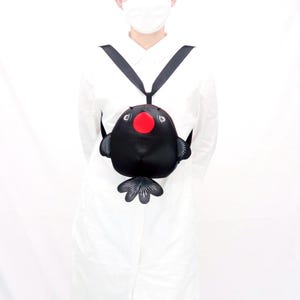 Puede incluir: Una mochila negra con forma de pez con una nariz roja y ojos blancos. La mochila tiene correas negras y un diseño en forma de aleta en la parte inferior. La mochila se lleva sobre los hombros.