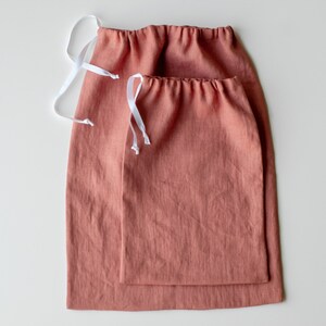 Puede incluir: Dos bolsas de cordón de lino de color terracota, una anidada dentro de la otra. Las bolsas tienen cordones de color blanco.