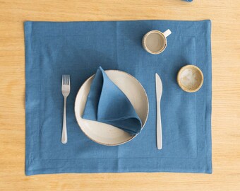 May include: A blue linen placemat with a white plate, a blue linen napkin, a fork, a knife, a small cup, and a small bowl on a wooden table.