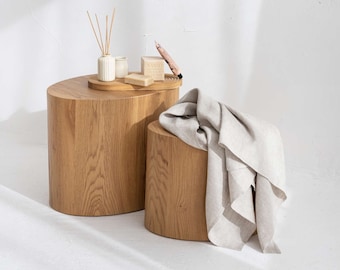 May include: Two wooden stools with a light brown finish. The larger stool has a tray on top with a small white ceramic diffuser, a small white ceramic candle, a bar of soap, and a wooden stick. A light gray linen throw is draped over the smaller stool.