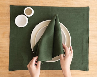 Könnte beinhalten: Eine grüne Leinen-Serviette, die zu einem Dreieck gefaltet ist, liegt auf einem weißen Teller. Der Teller liegt auf einem grünen Leinen-Platzdeckchen. Auf dem Platzdeckchen befinden sich zwei kleine weiße Schalen.