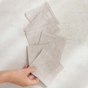 May include: A stack of six beige linen coasters, with one being held in a hand. The coasters are square and have a subtle texture.