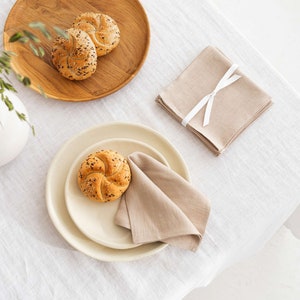 May include: A white linen tablecloth with a wooden tray holding two rolls with sesame seeds. A beige linen napkin is folded and placed on a beige plate with a roll on top.