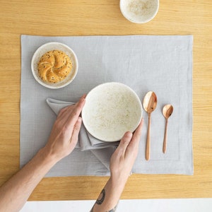 Może przedstawiać: Szara lniana serweta z białą, kropkowaną miską, małym talerzem z bułką i dwiema miedzianymi łyżkami. Miska jest trzymana przez osobę z wytatuowanym ramieniem.
