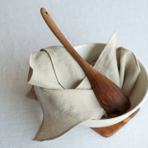 May include: A white ceramic bowl with a wooden spoon inside, both resting on a wooden coaster. The bowl is filled with a beige linen cloth.