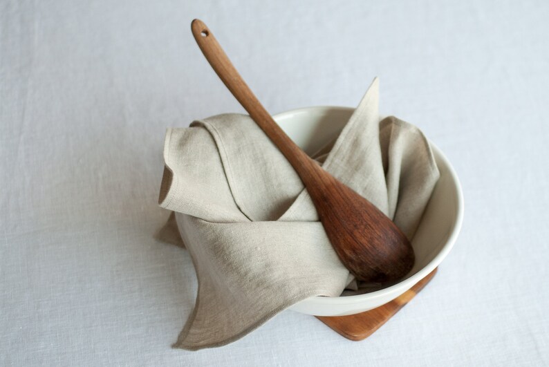 May include: A white ceramic bowl with a wooden spoon inside, both resting on a wooden coaster. The bowl is filled with a beige linen cloth.