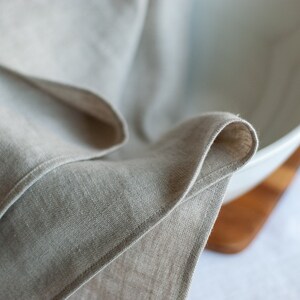 May include: A close-up of a folded linen kitchen towel draped over a white ceramic bowl. The bowl is sitting on a wooden surface.