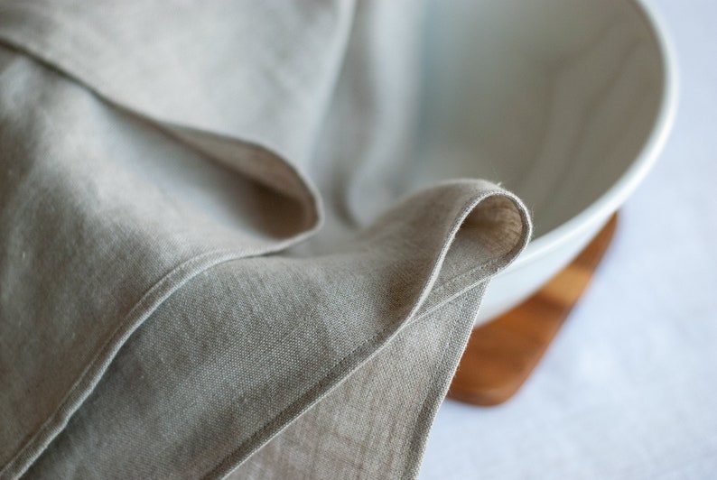 May include: A close-up of a folded linen kitchen towel draped over a white ceramic bowl. The bowl is sitting on a wooden surface.