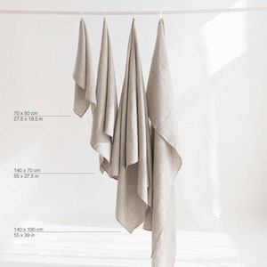 May include: Four linen towels of different sizes hanging on a clothesline. The towels are all a light beige color. The measurements of each towel are listed below the towels in inches and centimeters.