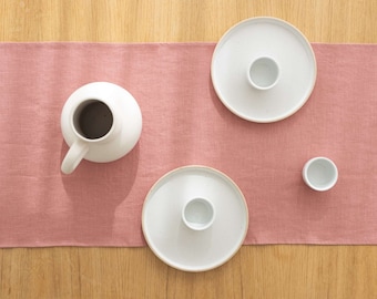 May include: A pink linen table runner with three white ceramic dishes and a white ceramic pitcher. The dishes have a small, white cup in the center of each.